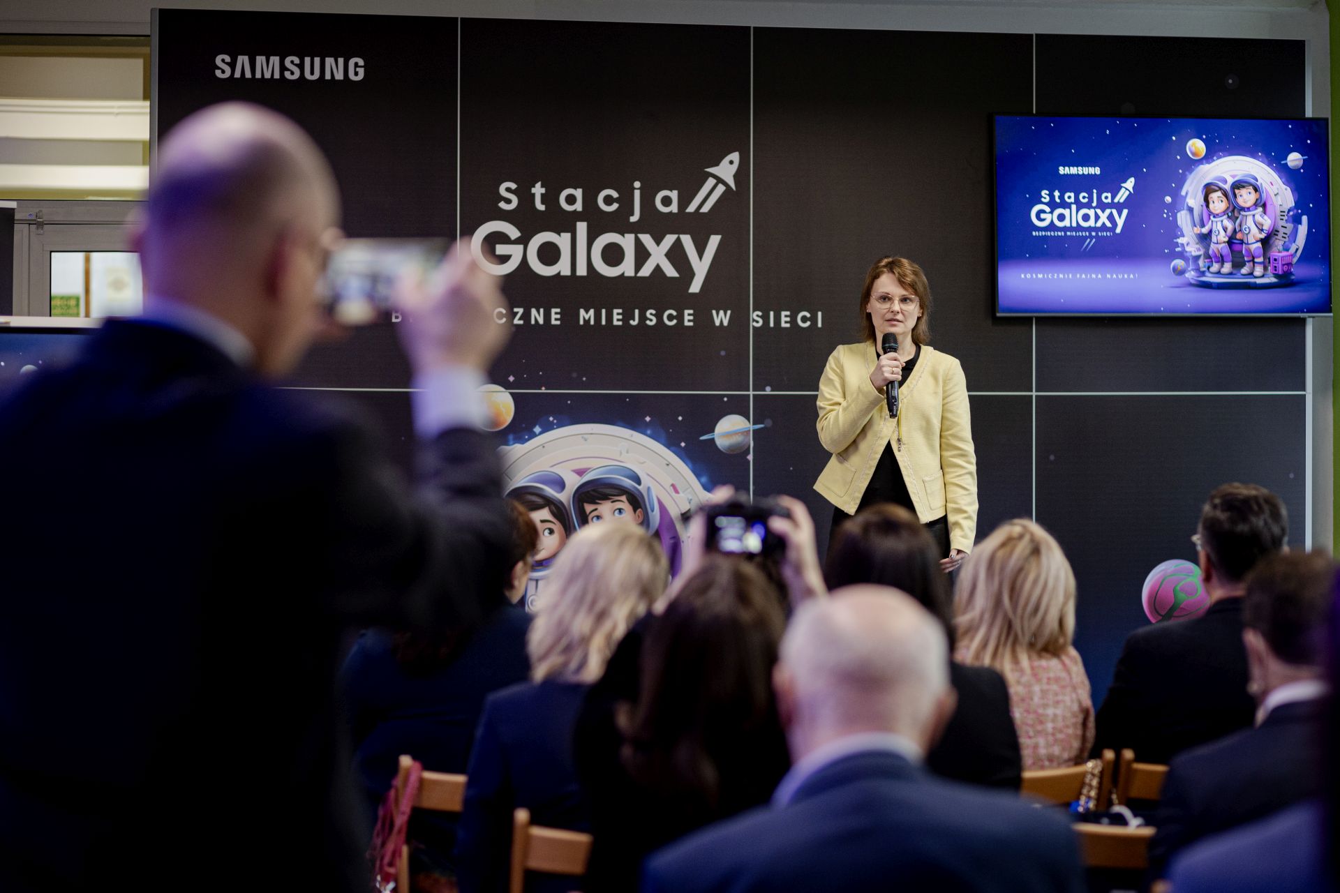 Zdjęcie przedstawiające grupę ludzi na sali. Na scenie elegancko ubrana kobieta z mikrofonem. Na pierwszym planie widownia. W tle ciemna ściana z napisem stacja galaxy.