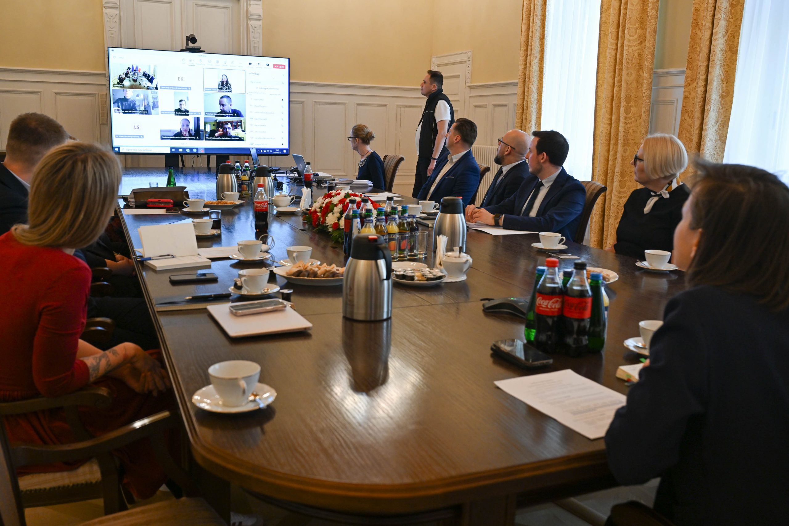Zdjęcie grupy ludzi przy dużym stole. Kobiety i mężczyźni są elegancko ubrani, patrzą się w stronę ekranu. Na ekranie połączeni uczestnicy spotkania.