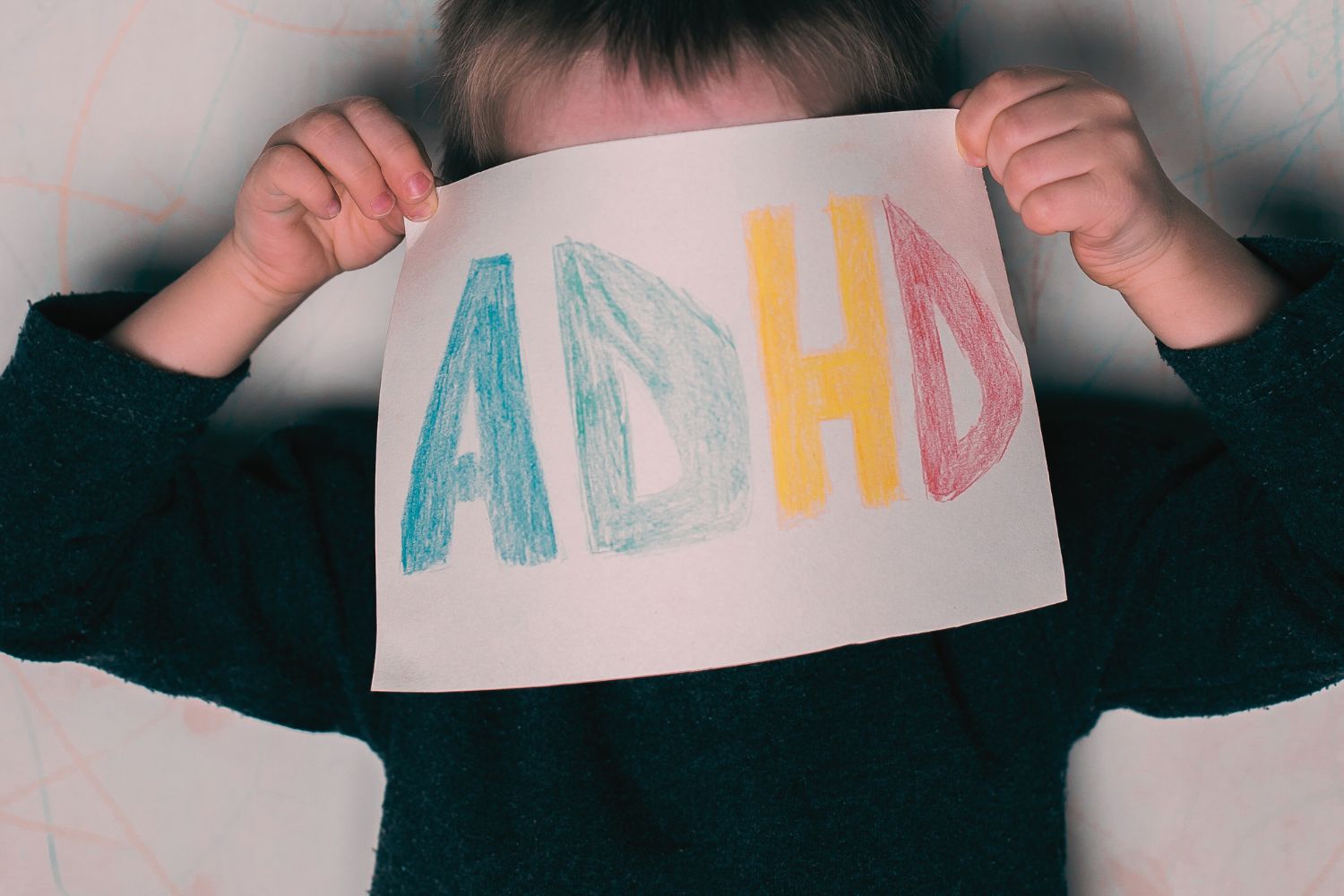 Na zdjęciu chłopiec, trzymający kartkę papieru z kolorowym napisem ADHD. Kartka zakrywa twarz chłopca