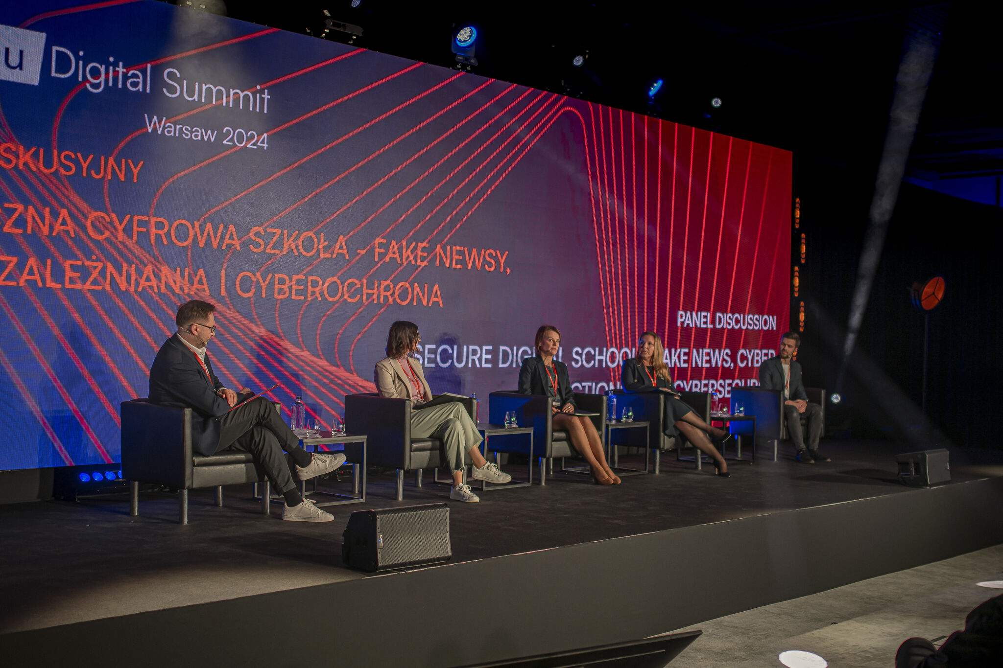 Zdjęcie przedstawiające ludzi na scenie. Siedzą na krzesłach, mają mikrofony. W tle ekran z wyświetlonym kolorowym obrazem i tekstem: Digital Summit, Warsaw 2024.