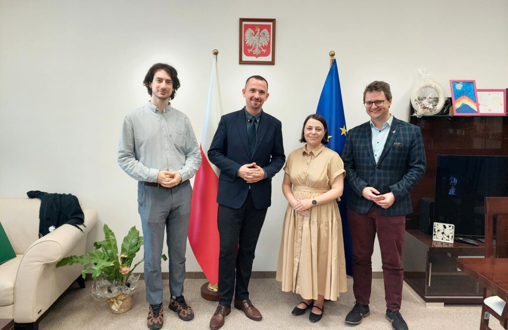 Zdjęcie elegancko ubranych osób - trzech mężczyzn i jednej kobiety. W tle flaga Polski i flaga Unii Europejskiej. Na jasnej ścianie godło Polski.