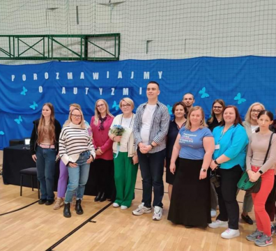 Zdjęcie grupy osób w sali sportowej. Za nimi niebiesko tło z napisem: porozmawiajmy o autyzmie.