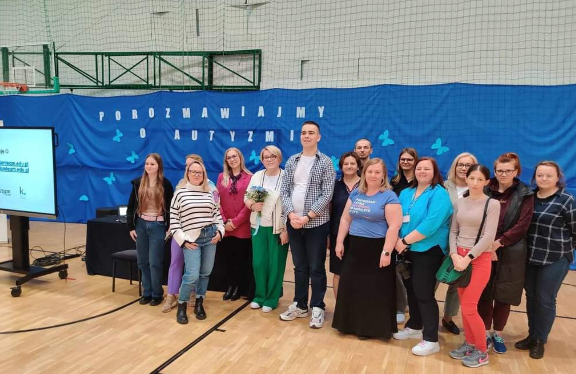 Zdjęcie grupy osób w sali sportowej. Za nimi niebiesko tło z napisem: porozmawiajmy o autyzmie.