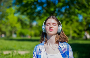Zdjęcie nastolatki ze słuchawkami na uszach. Ma ciemne, średniej długości włosy. Jest ubrana w niebieską koszulę. W tle zielone drzewa.